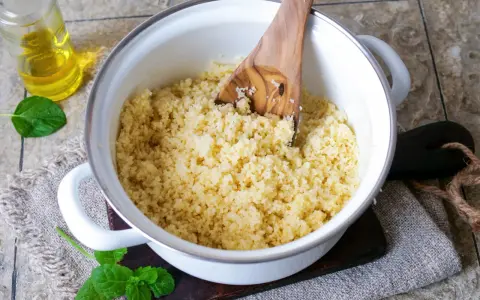 Frisch zubereiteter Couscous in einem Topf, bereit zum Servieren. Ein Holzlöffel lockert die Körner auf. Daneben eine Flasche Öl und Minzblätter.