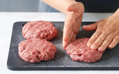 Hände formen rohe Burger-Patties auf einem dunklen Schneidebrett.