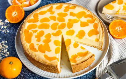 Ein saftiger faule weiber kuchen mit Mandarinenstücken, der auf einem Teller liegt. Daneben liegen frische Mandarinen und eine Tortenschaufel.