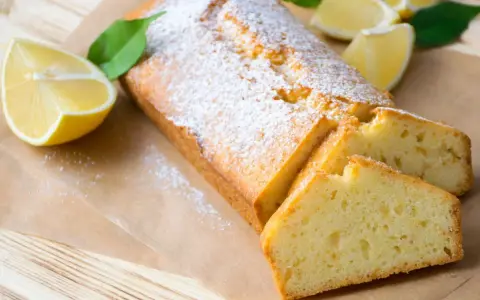 Saftiger veganer Zitronenkuchen, bestäubt mit Puderzucker, neben frischen Zitronenspalten und grünen Blättern.