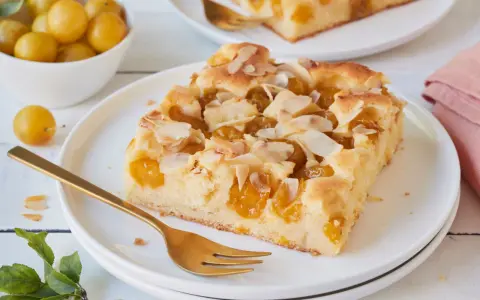 Ein Stück saftiger Mirabellenkuchen mit Mandelblättchen, serviert auf einem Teller mit einer goldenen Gabel. Daneben eine Schale voller frischer Mirabellen.