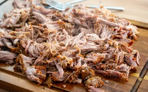 Saftiges pulled pork, zart zerfallen, liegt auf einem Holzbrett. Eine Gabel liegt bereit, um das Fleisch zu servieren.