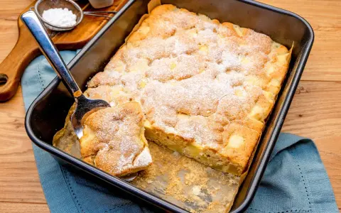 Ein Stück vom schneller saftiger apfelkuchen vom blech wird mit einem Kuchenheber herausgehoben. Mit Puderzucker bestäubt.