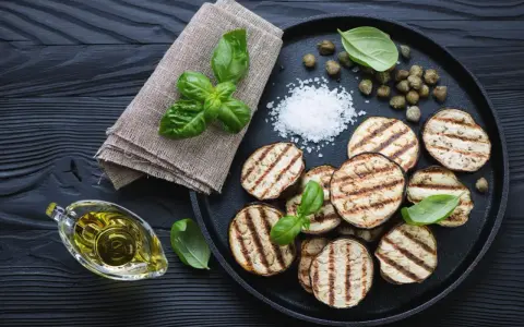 Gegrillte Auberginenscheiben mit Basilikum, Kapern und grobem Salz auf schwarzem Holz. Perfekt zum Aubergine grillen.