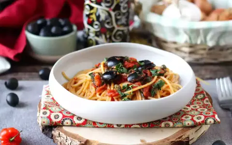 Eine Schüssel Spaghetti Puttanesca, garniert mit schwarzen Oliven und Kräutern, auf einem rustikalen Holzständer.