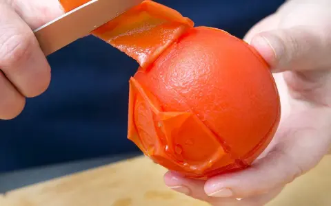 Hände ziehen mit einem Messer die Haut von einer Tomate ab. Die Tomate ist fast komplett geschält.
