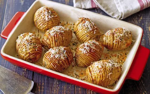 A red baking dish holds golden-brown hasselback kartoffeln, sprinkled with grated cheese and herbs.