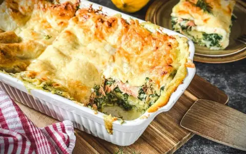 Herzhafte Lachs Spinat Lasagne, goldbraun überbacken, dampft in einer weißen Auflaufform. Ein Stück auf einem Teller zeigt die Schichten.