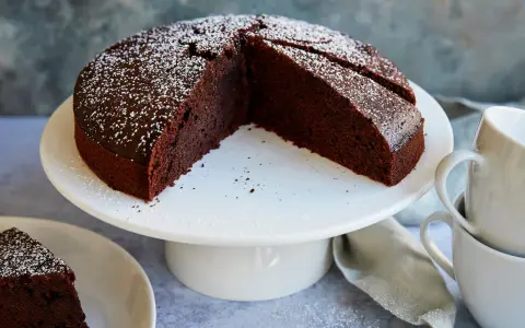 Ein saftiger veganer Schokokuchen, bestäubt mit Puderzucker, wartet auf Genuss. Daneben liegen zwei Tassen.