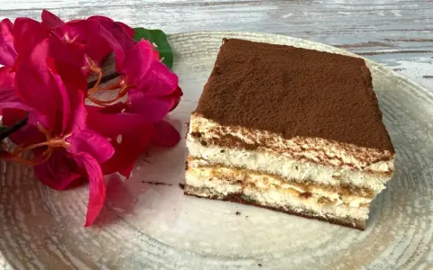 Ein Stück Tiramisu, perfekt für Ihr nächstes tiramisu rezept, liegt auf einem Teller neben leuchtend roten Blüten.