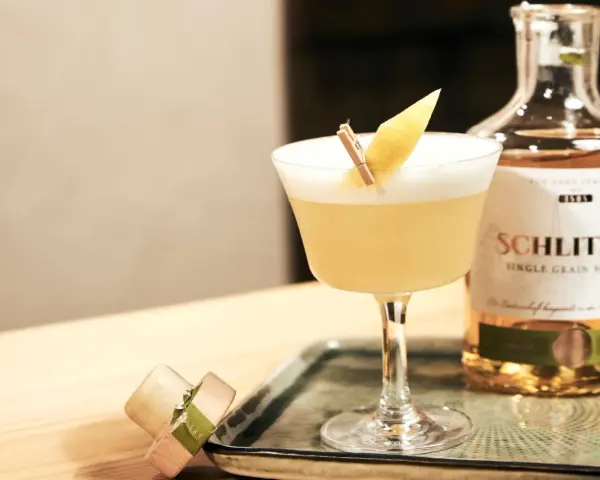 Ein erfrischender Sour Cocktail mit Zitronenspalte, neben einer Flasche Schlitzer Single Grain Whisky. Perfekt für ein Sour Cocktail Rezept.