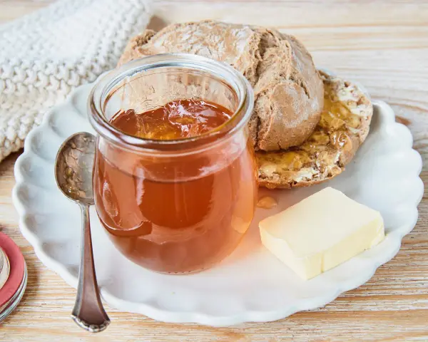 Glas mit goldbraunem Apfelgelee aus Apfelsaft, daneben ein Stück Butter und ein halbes Brot auf einem Teller.