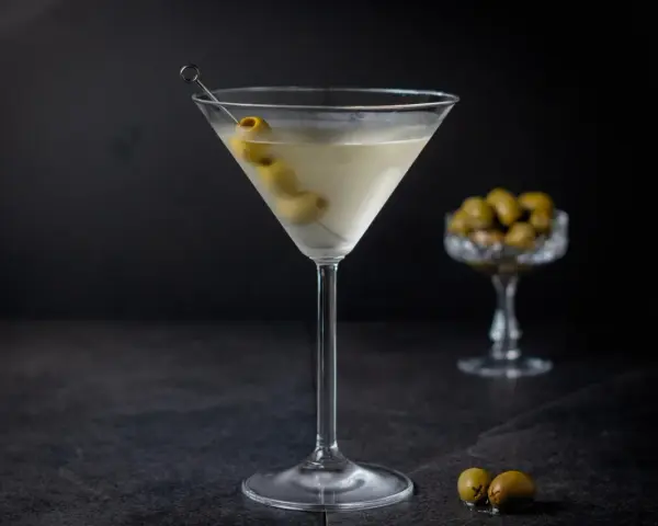 Ein klassischer martini trocken, garniert mit Oliven, steht auf einem dunklen Tisch. Im Hintergrund eine Schale mit weiteren Oliven.