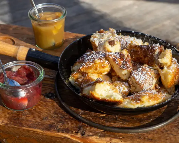 Ein frisch zubereiteter Kaiserschmarrn im Pfännchen, bestäubt mit Puderzucker, daneben Apfelmus und Zwetschgenröster. Ein leckeres Kaiserschmarrn Rezept zum Nachmachen.