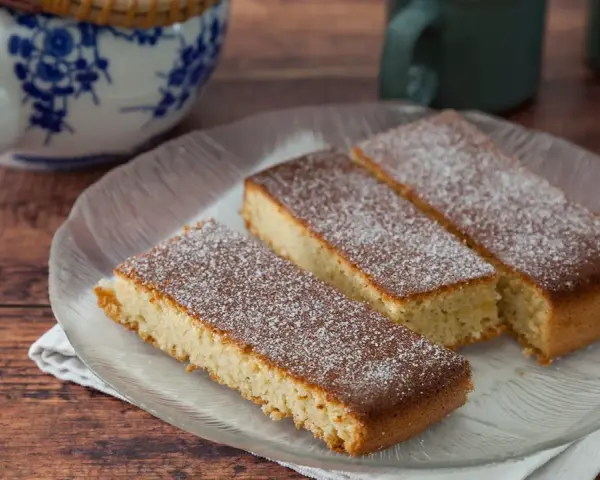 Drei rechteckige Stücke mandelkuchen vom blech, mit Puderzucker bestäubt, auf einem Glas-Teller.