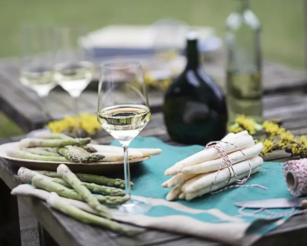 Ein Glas Weißwein steht neben frischem Spargel auf einem Holztisch. Die Frage welcher Wein zum Spargel passt, wird hier visuell beantwortet.