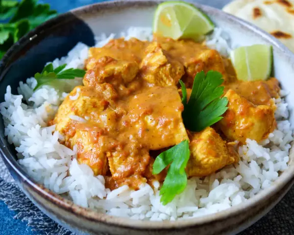 Ein köstliches indisches Curry mit zarten Hähnchenstücken in cremiger Sauce, serviert auf lockerem Reis, garniert mit frischer Petersilie und Limettenspalten.