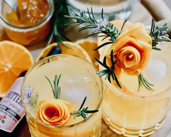 Zwei erfrischende Cocktails mit Orangenscheiben, Rosmarin und einer Orangenschalenrose. Daneben eine Flasche "Bitters" mit Lavendelgeschmack.