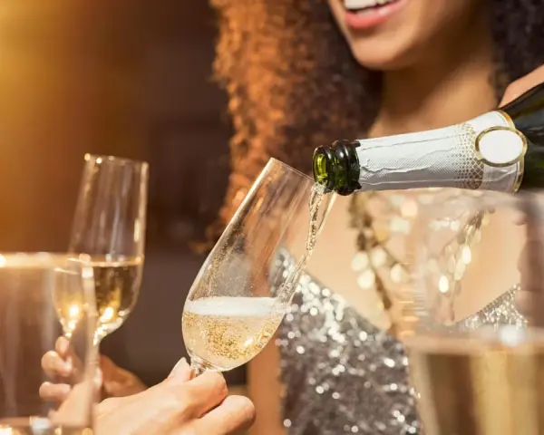 Sekt wird in ein Glas gegossen, während Freunde anstoßen. Der Unterschied zwischen Wein und Sekt ist die Kohlensäure, die Sekt prickelnd macht.
