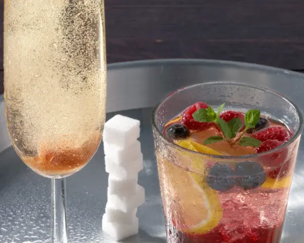 Sektglas mit sprudelndem Champagner-Cocktail, daneben ein Glas mit Beeren, Zitrone und Minze. Ein Stapel Zuckerwürfel.