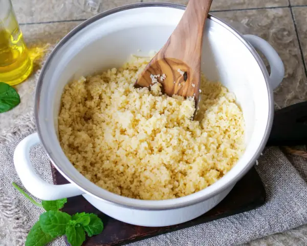 Frisch zubereiteter Couscous in einem Topf, bereit zum Servieren. Ein Holzlöffel lockert die Körner auf. Daneben eine Flasche Öl und Minzblätter.