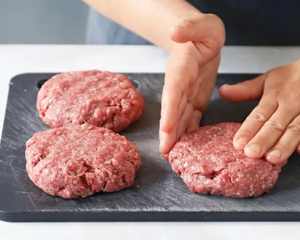 Hände formen rohe Burger-Patties auf einem dunklen Schneidebrett.