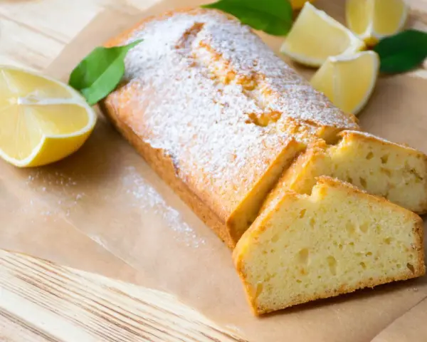 Saftiger veganer Zitronenkuchen, bestäubt mit Puderzucker, neben frischen Zitronenspalten und grünen Blättern.