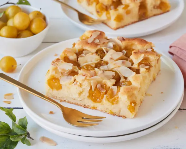 Ein Stück saftiger Mirabellenkuchen mit Mandelblättchen, serviert auf einem Teller mit einer goldenen Gabel. Daneben eine Schale voller frischer Mirabellen.