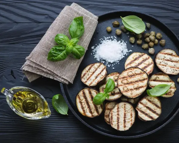 Gegrillte Auberginenscheiben mit Basilikum, Kapern und grobem Salz auf schwarzem Holz. Perfekt zum Aubergine grillen.