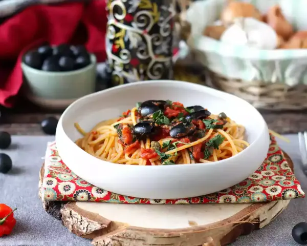 Eine Schüssel Spaghetti Puttanesca, garniert mit schwarzen Oliven und Kräutern, auf einem rustikalen Holzständer.