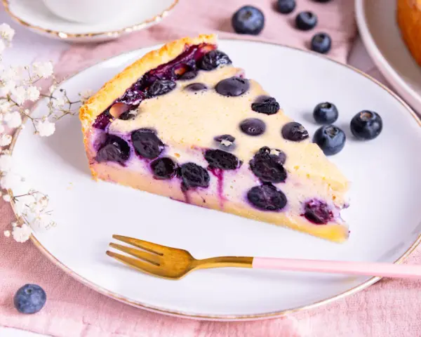 Ein Stück saftiger Heidelbeerkuchen Landfrauenart auf einem Teller, garniert mit frischen Beeren und einer Gabel.