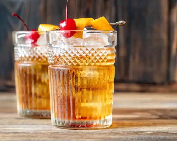 Zwei klassische Gin Old Fashioned Cocktails, garniert mit Kirschen und Orangenzesten, auf einem Holztisch.