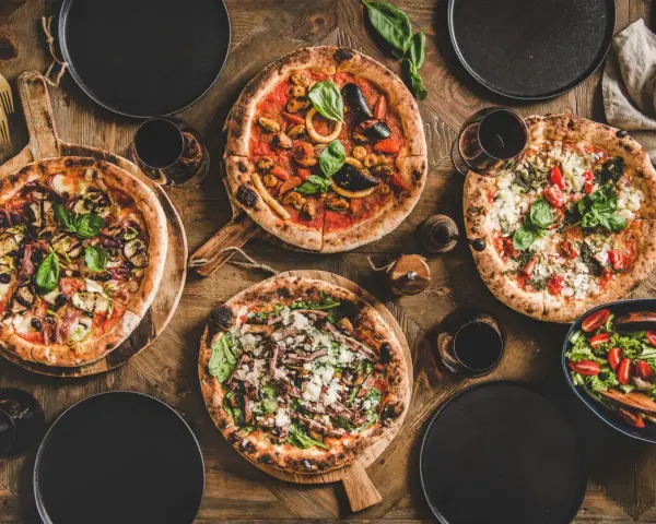 Vier leckere Pizzen und ein Salat auf Holztisch. Die Frage welcher Wein zu Pizza passt, wird hier mit Rotwein beantwortet.