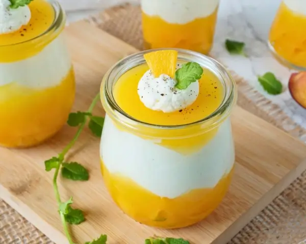 Erfrischendes Solero Dessert im Glas mit Mango-Fruchtsoße, weißer Creme und einem Klecks Sahne, garniert mit Minze und Orangenspalte.