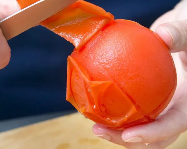 Hände ziehen mit einem Messer die Haut von einer Tomate ab. Die Tomate ist fast komplett geschält.