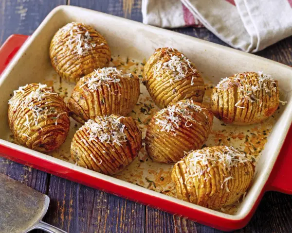 A red baking dish holds golden-brown hasselback kartoffeln, sprinkled with grated cheese and herbs.