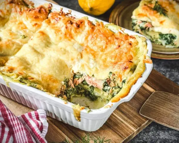Herzhafte Lachs Spinat Lasagne, goldbraun überbacken, dampft in einer weißen Auflaufform. Ein Stück auf einem Teller zeigt die Schichten.
