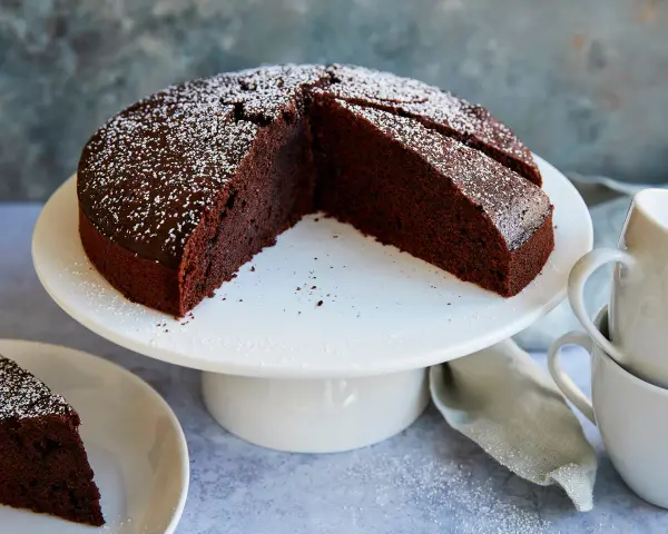 Ein saftiger veganer Schokokuchen, bestäubt mit Puderzucker, wartet auf Genuss. Daneben liegen zwei Tassen.