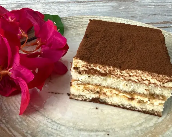 Ein Stück Tiramisu, perfekt für Ihr nächstes tiramisu rezept, liegt auf einem Teller neben leuchtend roten Blüten.