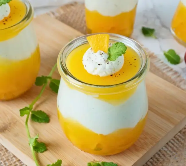 Erfrischendes Solero Dessert im Glas mit Mango-Fruchtsoße, weißer Creme und einem Klecks Sahne, garniert mit Minze und Orangenspalte.