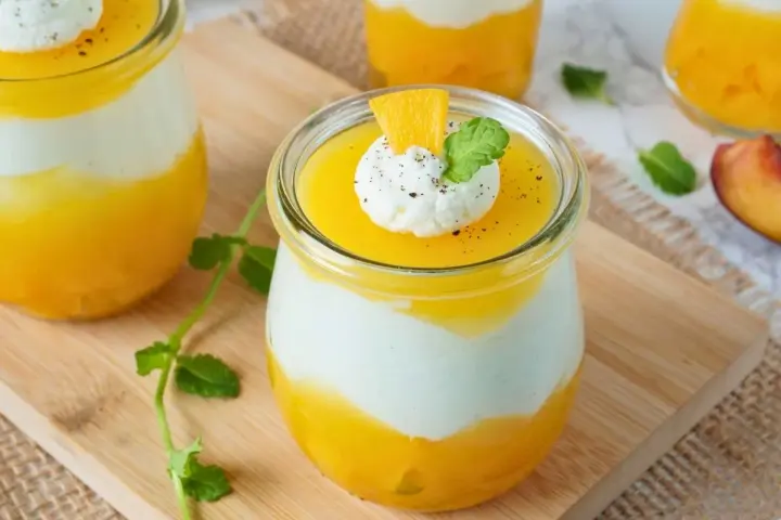 Erfrischendes Solero Dessert im Glas mit Mango-Fruchtsoße, weißer Creme und einem Klecks Sahne, garniert mit Minze und Orangenspalte.
