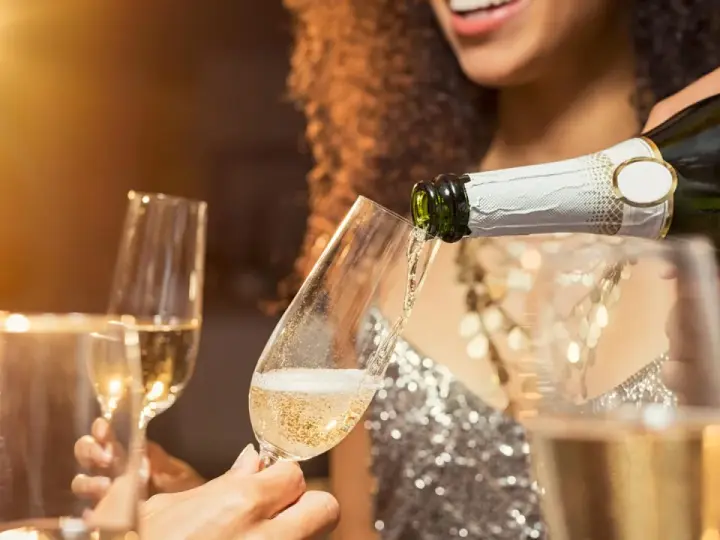 Sekt wird in ein Glas gegossen, während Freunde anstoßen. Der Unterschied zwischen Wein und Sekt ist die Kohlensäure, die Sekt prickelnd macht.