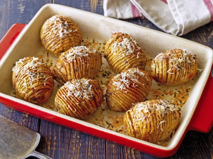 A red baking dish holds golden-brown hasselback kartoffeln, sprinkled with grated cheese and herbs.