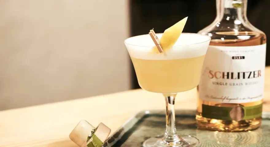 Ein erfrischender Sour Cocktail mit Zitronenspalte, neben einer Flasche Schlitzer Single Grain Whisky. Perfekt für ein Sour Cocktail Rezept.