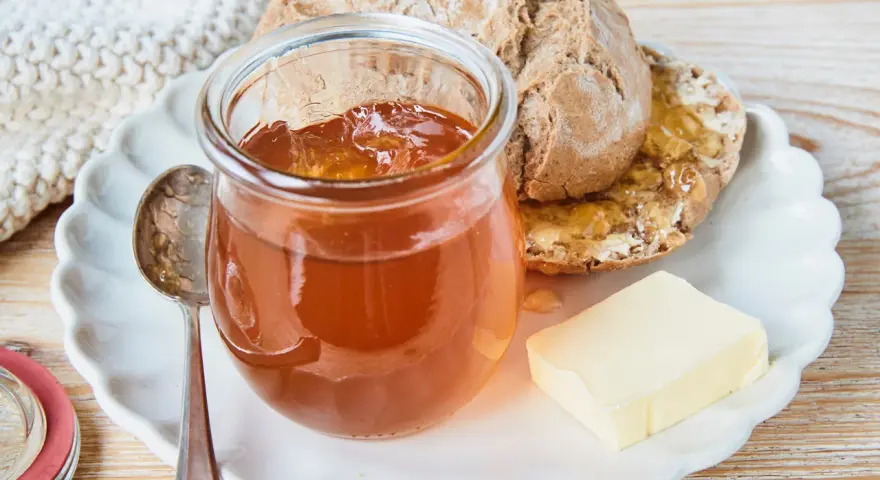 Glas mit goldbraunem Apfelgelee aus Apfelsaft, daneben ein Stück Butter und ein halbes Brot auf einem Teller.