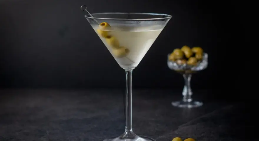 Ein klassischer martini trocken, garniert mit Oliven, steht auf einem dunklen Tisch. Im Hintergrund eine Schale mit weiteren Oliven.