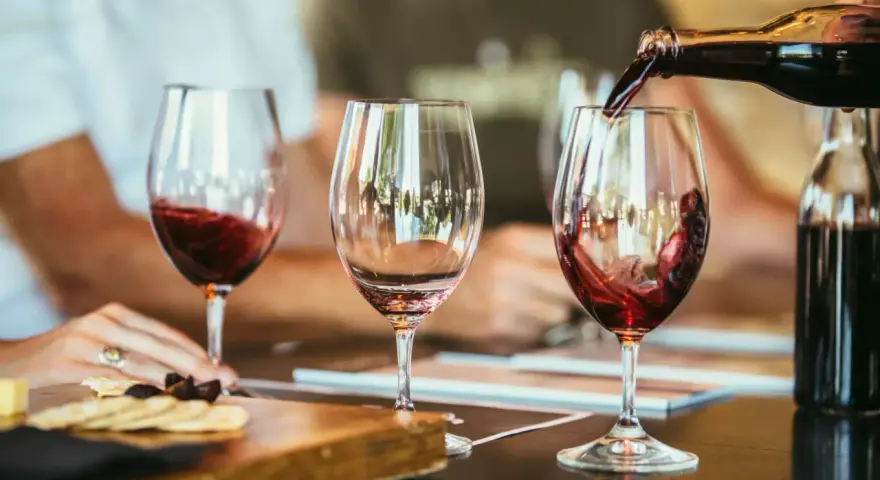 Rotwein wird in ein Glas gegossen, der Duft von **Wein** und **Tanninen** liegt in der Luft. Daneben ein leeres Glas und ein Teller mit Käse und Crackern.