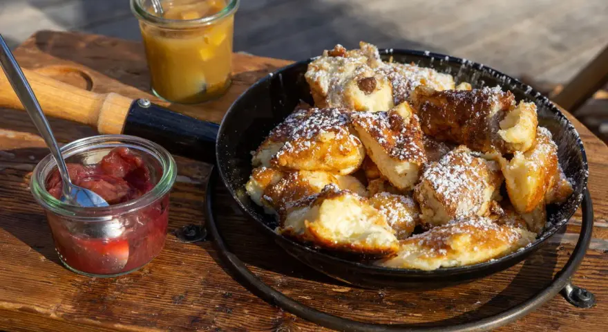 Ein frisch zubereiteter Kaiserschmarrn im Pfännchen, bestäubt mit Puderzucker, daneben Apfelmus und Zwetschgenröster. Ein leckeres Kaiserschmarrn Rezept zum Nachmachen.