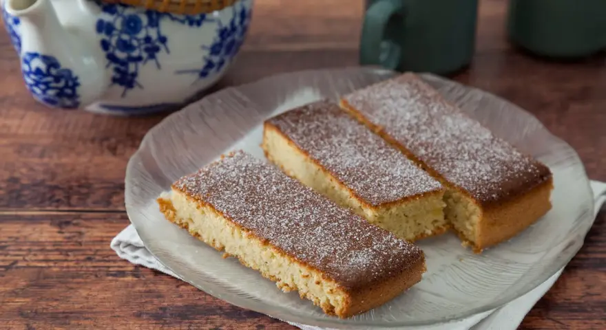 Drei rechteckige Stücke mandelkuchen vom blech, mit Puderzucker bestäubt, auf einem Glas-Teller.