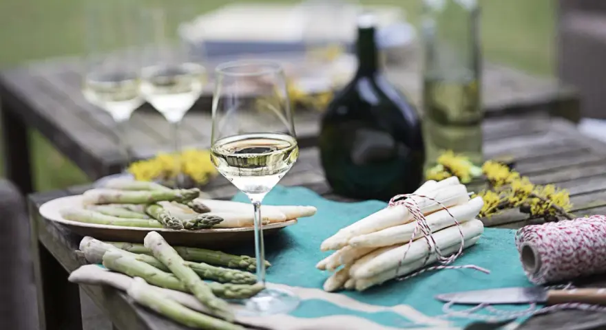 Ein Glas Weißwein steht neben frischem Spargel auf einem Holztisch. Die Frage welcher Wein zum Spargel passt, wird hier visuell beantwortet.
